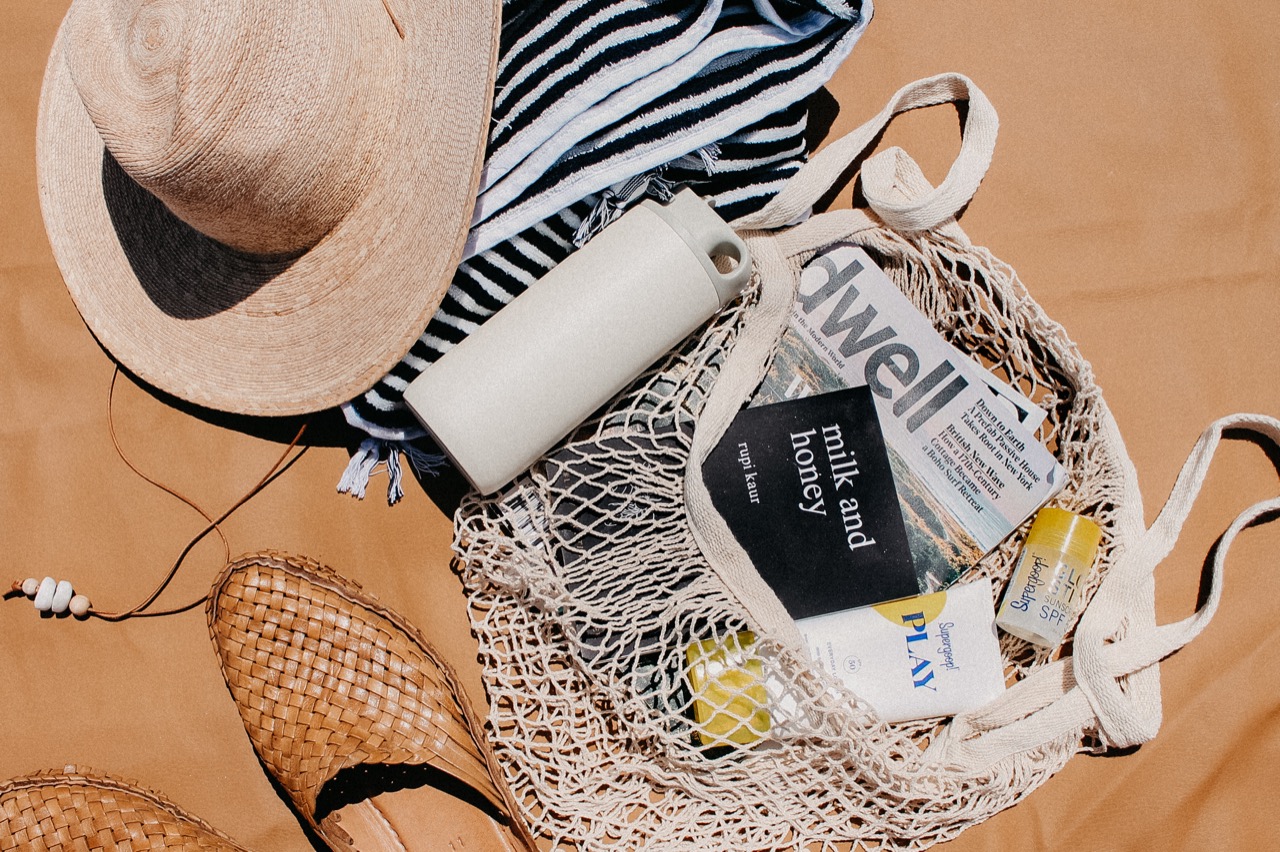 Overhead image of a tan beach blanket with hat, towel, woven shoes, water bottle, market bag, book, magazines, and sunscreen.