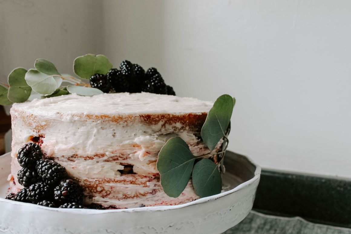 Blackberry lavender cake on gray cake stand with gray linens beneath and blackberry and eucalyptus decoration on cake.
