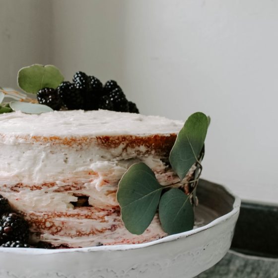 Blackberry lavender cake on gray cake stand with gray linens beneath and blackberry and eucalyptus decoration on cake.