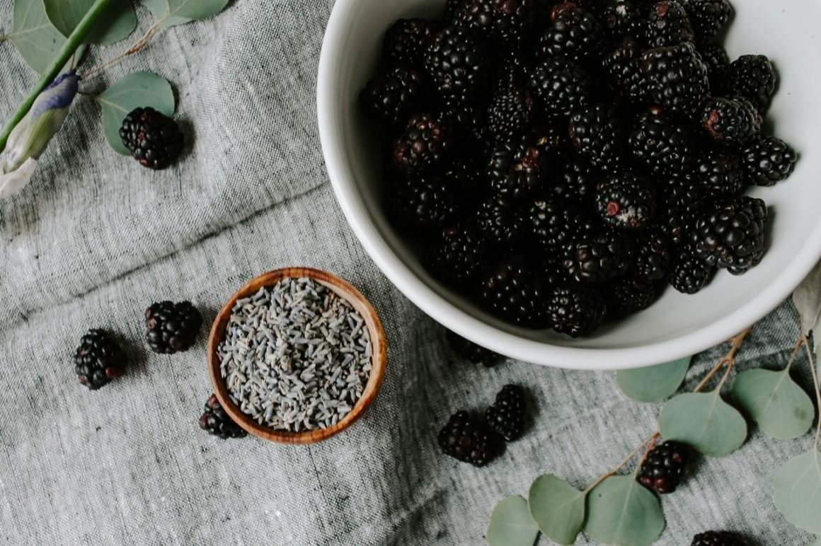 Overhead image of a bowl of blackberries and a wooden bowl of lavender on gray linens with extra blackberries and eucalyptus on linens.