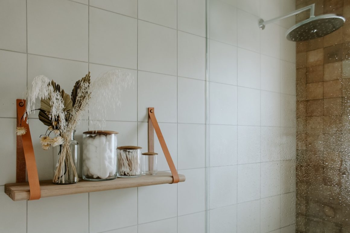 Square tiled wall of bathroom with hanging wooden shelf holding various jars and a vase of dried flowers which hangs next to glass wall of shower.