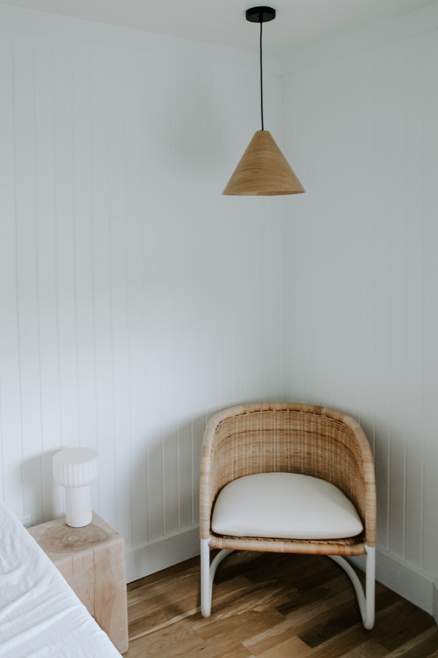 Corner of bedroom with a ratan chair with white cushion sitting under a wooden pendant lamp with white wood paneled walls.