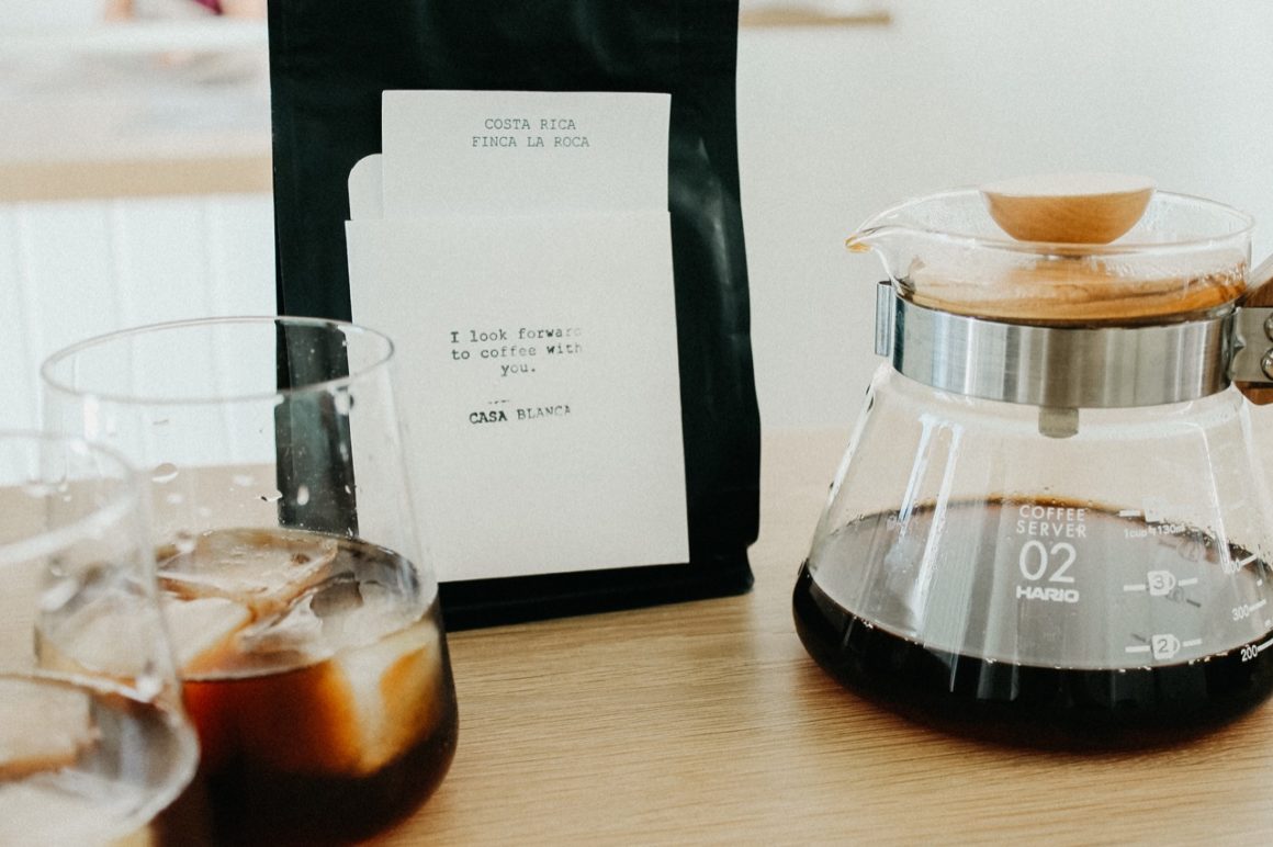 Bag of coffee with two glasses of iced coffee and a coffee pourer in the foreground.
