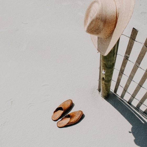 Woven shoes laying on flat sand with straw hat propped up on sand dune posts.