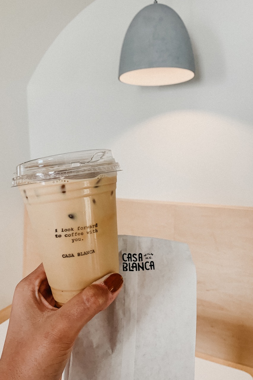 Person's hand holding an iced latte with a paper bag with seating booth in background.
